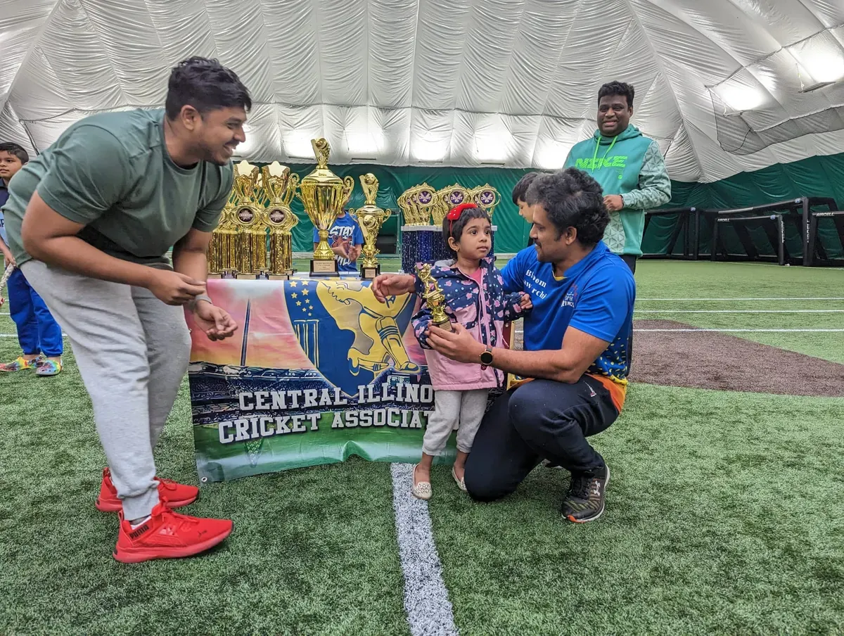 Adults and children gathered beside a table of cricket trophies indoors.